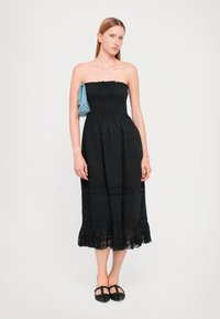 Vestido preto sem alças com um tecido texturizado e uma bainha de renda em camadas. Uma bolsa em padrão xadrez azul claro está pendurada em um ombro.