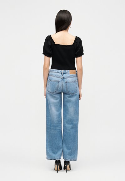 Kvinne med langt mørkt hår iført svart topp med pufferm, lyseblå vide jeans og svarte høye hæler, står med ryggen til.