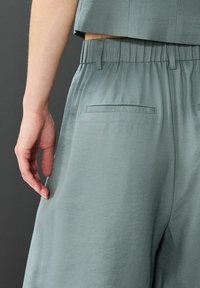 Světle šedé kalhoty s širokými nohavicemi a elastickým pasem, se dvěma zadními kapsami a hladkým texturovaným materiálem.