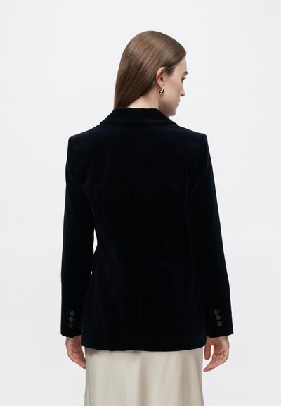 Blazer in velluto nero con vestibilità su misura, rever a lancia e polsini con bottoni. Texture liscia, design classico, cuciture visibili e due spaccature posteriori.