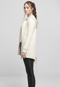 Sudadera con cremallera de color beige claro, con capucha, mangas largas, aberturas laterales y detalles de cordón. Combinada con leggings negros con rayas verticales.