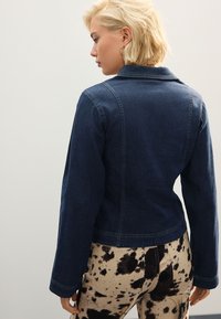 Chaqueta de mezclilla oscura con un diseño estructurado, que cuenta con mangas largas y una textura suave. Combinada con pantalones beige adornados con manchas negras.