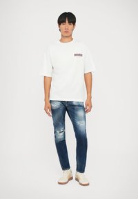 Camiseta blanca de manga corta con el logo "DSQUARED2", combinada con jeans rasgados de color azul oscuro y zapatillas beige con suelas marrones.