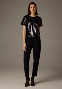 Schwarzes Paillettenshirt mit kurzen Ärmeln, kombiniert mit schwarzen Hosen mit Kordelzug. Das Model trägt schwarze Spitzenknöchelsocken und schwarze Schuhe mit spitzer Zehenpartie.