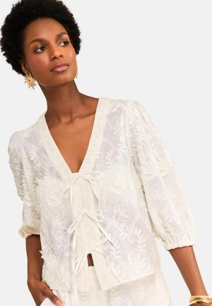 Femme aux cheveux courts et bouclés portant un chemisier blanc brodé de fleurs avec des fermetures à nouer et des boucles d'oreilles dorées en forme d'éclat de soleil.