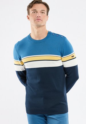 Homme portant un pull rayé bleu, jaune et blanc avec des boutons sur l'épaule, debout les mains derrière le dos sur fond blanc.
