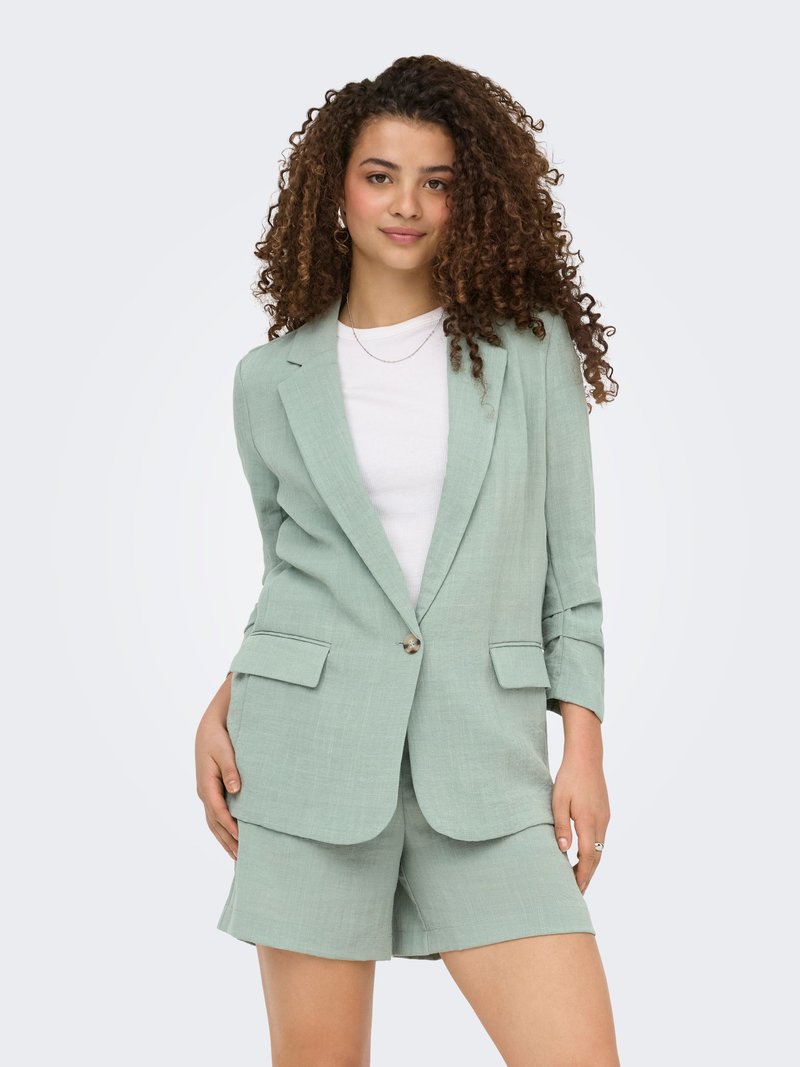 Jeune femme aux cheveux bouclés porte un blazer vert clair et un short assorti sur un t-shirt blanc, debout devant un fond uni.