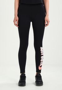Beine in schwarzen Nike-Leggings mit pinkem "Just Do It"-Text am linken Bein und schwarzen Sneakers, die auf einem weißen Boden stehen.