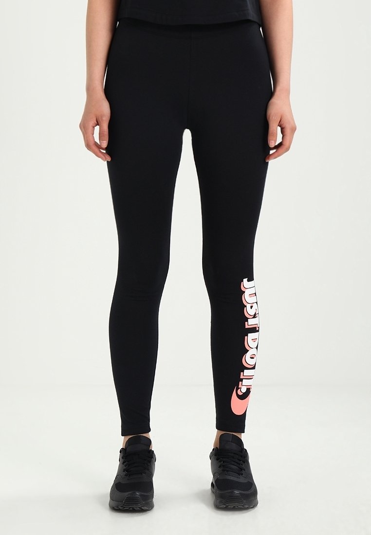 Beine in schwarzen Nike-Leggings mit pinkem "Just Do It"-Text am linken Bein und schwarzen Sneakers, die auf einem weißen Boden stehen.