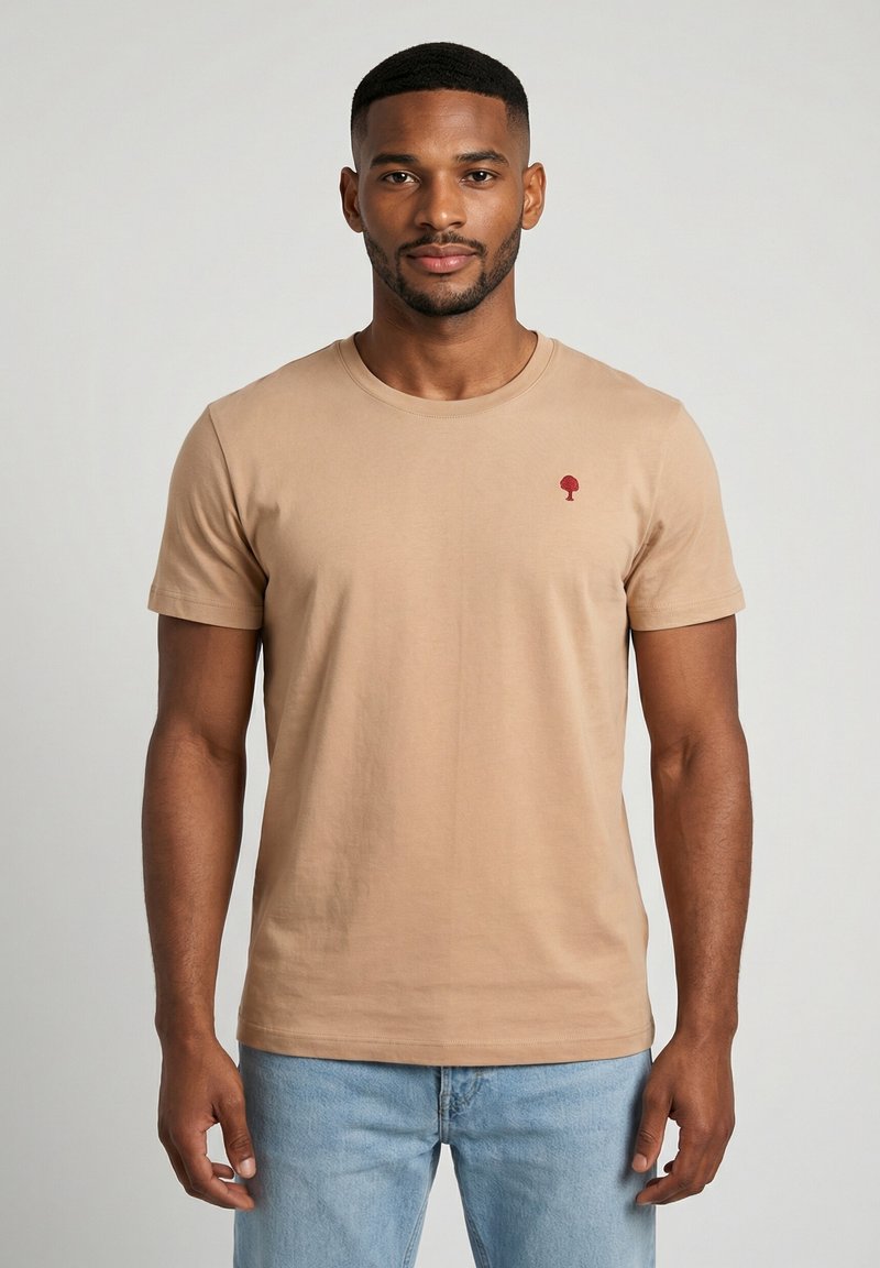 Uomo con capelli corti e barba che indossa una maglietta beige con un piccolo logo rosso a forma di albero e jeans azzurri, in piedi contro uno sfondo grigio.