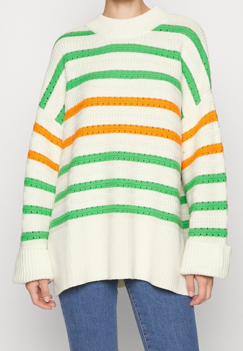 Personne portant un pull en maille blanc oversize avec des rayures horizontales vertes et orange et un jean bleu, debout devant un fond uni.