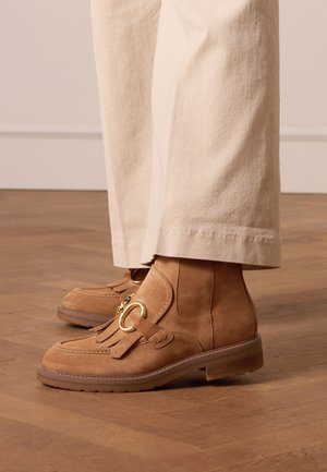 Tan suede ankle boots with fringe detail, gold hardware, and a round accent. Flat rubber sole and stitched outline, paired with light trousers.