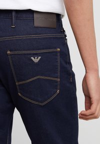 Närbild av en bakficka på mörkblå jeans med Emporio Armani-logotyp och en brun skinnmärke på midjebandet, buren av en person i vit skjorta.