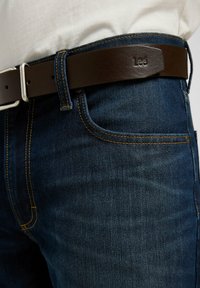 Mörkblå denimjeans med kontrasterande gul sömnad, prydda med ett brunt läderbälte med en silverfärgad metalklämma och ett präglat logotyp.