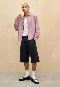 Camisa de botão às riscas rosa, t-shirt branca por baixo, calções pretos, meias brancas e sapatos pretos. Design simples e corte descontraído.