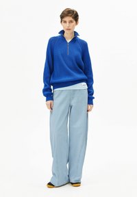 Blauer gestrickter Halb-Reißverschluss-Pullover mit geripptem Muster, kombiniert mit hellblauen Jeans mit weitem Bein und kontrastierenden Nähten. Schwarze und gelbe Schuhe.