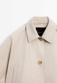 Trench-coat beige avec col pointu, deux boutons marron et poche latérale, marque Massimo Dutti.