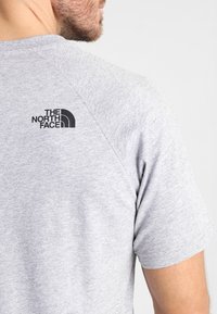 Camiseta gris de manga corta con cuello redondo. Presenta un logo negro "THE NORTH FACE" en la parte superior de la espalda. Textura suave, similar al algodón.