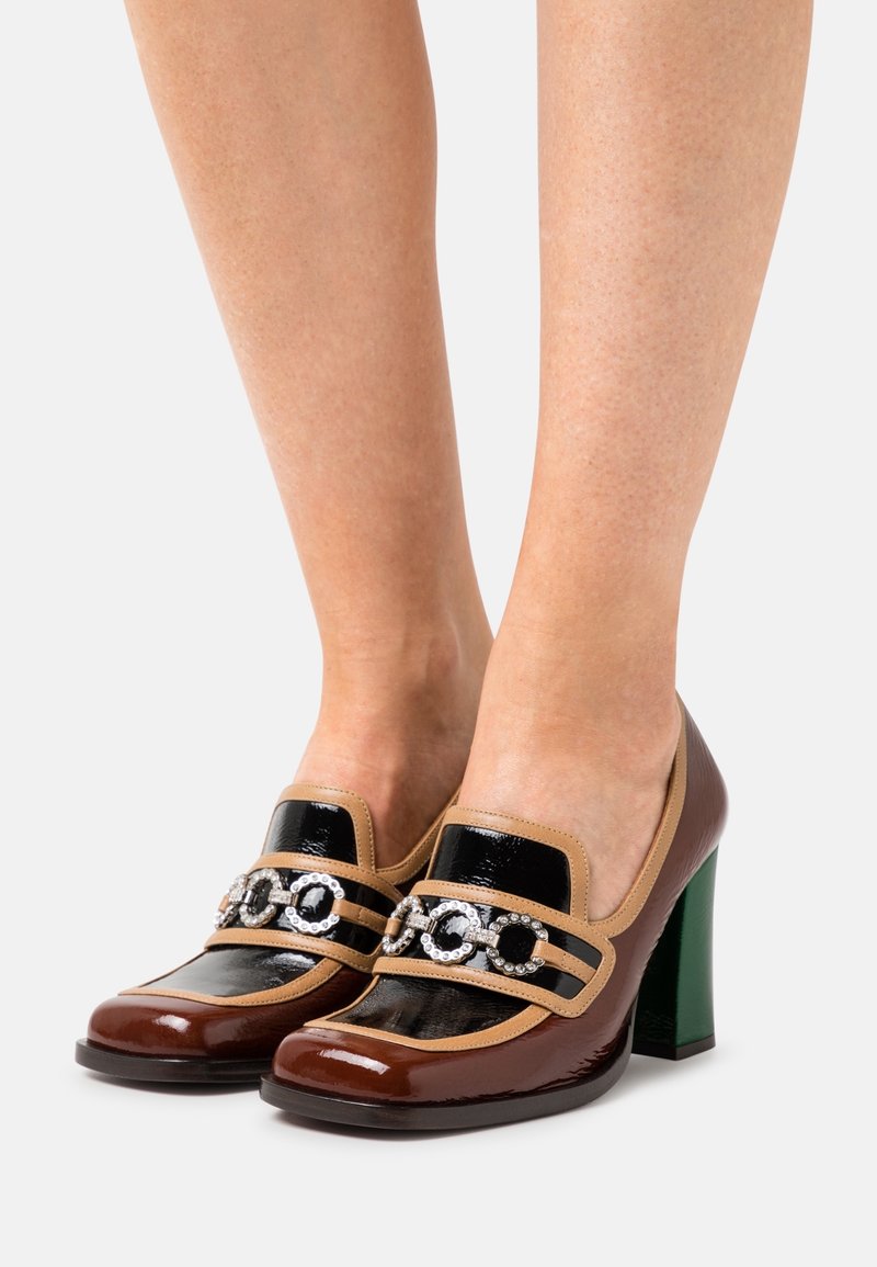 Chaussures en cuir verni marron et noir avec une bande beige et des éléments décoratifs. Dotées d'un talon bloc vert et d'une texture brillante.