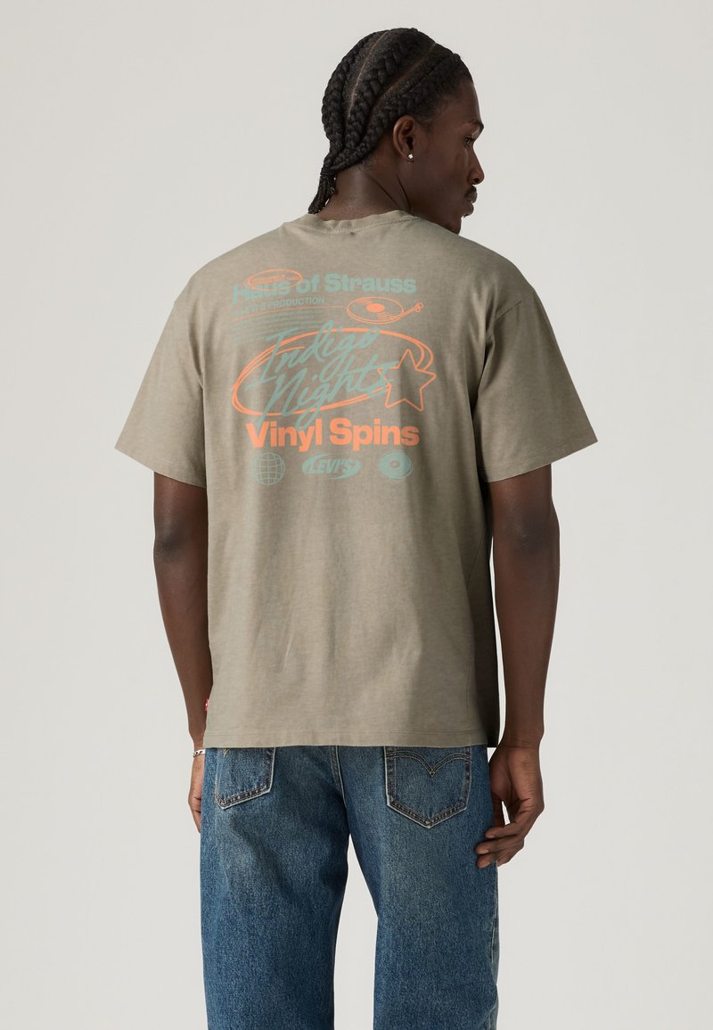 Homme aux cheveux tressés portant un t-shirt graphique gris avec l'inscription « Indigo Nights Vinyl Spins » et un jean bleu, tournant le dos sur un fond uni.