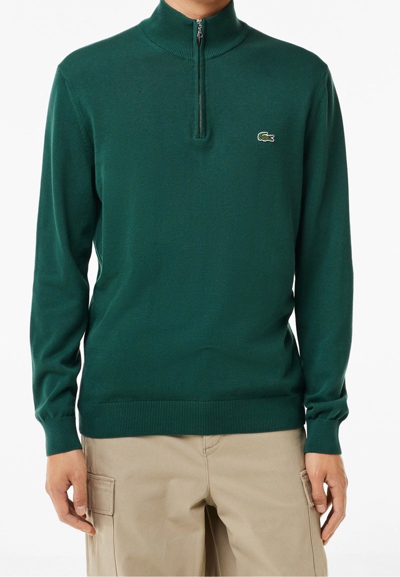 Homme portant un pull à col montant zippé vert foncé avec un petit logo crocodile sur la poitrine et un pantalon cargo beige.