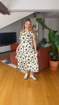 Ung kvinne i blomstrete kjole og joggesko står innendørs nær en sofa og en stor potteplante, holder kjolekanten med begge hender.