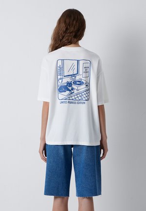Person with long hair wearing a white t-shirt with a blue cat and record player illustration and blue denim shorts, seen from the back.