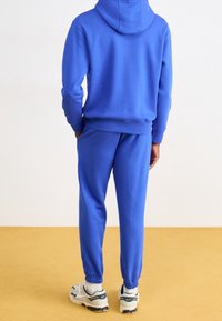 Blå bomullshuvtröja med känguruficka och matchande joggers, med elastiska muddar och midja, kombinerad med vita och grå sneakers.