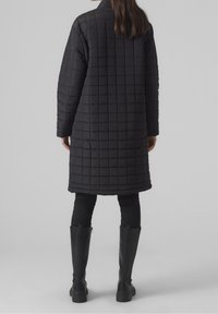 Abrigo largo acolchado negro con un corte relajado. Presenta un cuello alto, costuras de hombro estructuradas y bolsillos laterales. Combinado con leggings negros y botas altas.