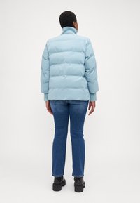 Chaqueta acolchada de color azul claro con cuello alto, que presenta un patrón de costura horizontal y mangas largas, combinada con jeans de color azul oscuro y botas de tobillo.