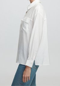 Chemise blanche à boutons, coupe décontractée, avec deux poches poitrine et manches longues. Le tissu semble lisse avec un léger éclat.