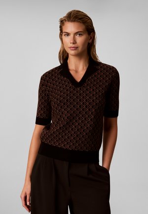 Femme aux cheveux blonds mouillés portant un haut en maille marron à manches courtes avec un col noir et un pantalon noir, posant avec une main dans la poche.