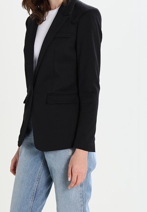 Femme portant un blazer noir cintré sur un top blanc côtelé, associé à un jean denim bleu clair, debout devant un fond blanc uni.