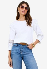 Haut blanc à manches longues avec des accents en dentelle sur les manches, associé à un jean bleu clair et une ceinture marron. Le mannequin porte des lunettes de soleil surdimensionnées.