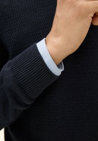 Granatowy sweter o fakturowanym splocie, z prążkowanymi mankietami. Pod rękawem swetra widoczna jest jasnoniebieska koszula w kratę.