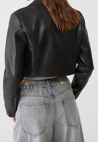 Giacca cropped in pelle nera con spalle strutturate. Indossata sopra pantaloni grigi a vita alta con fascia elastica in vita e tasche posteriori.