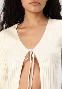 Cream ribbed knit top with a deep V-neck and front tie closure. The fabric is textured and fitted, accentuating the silhouette.