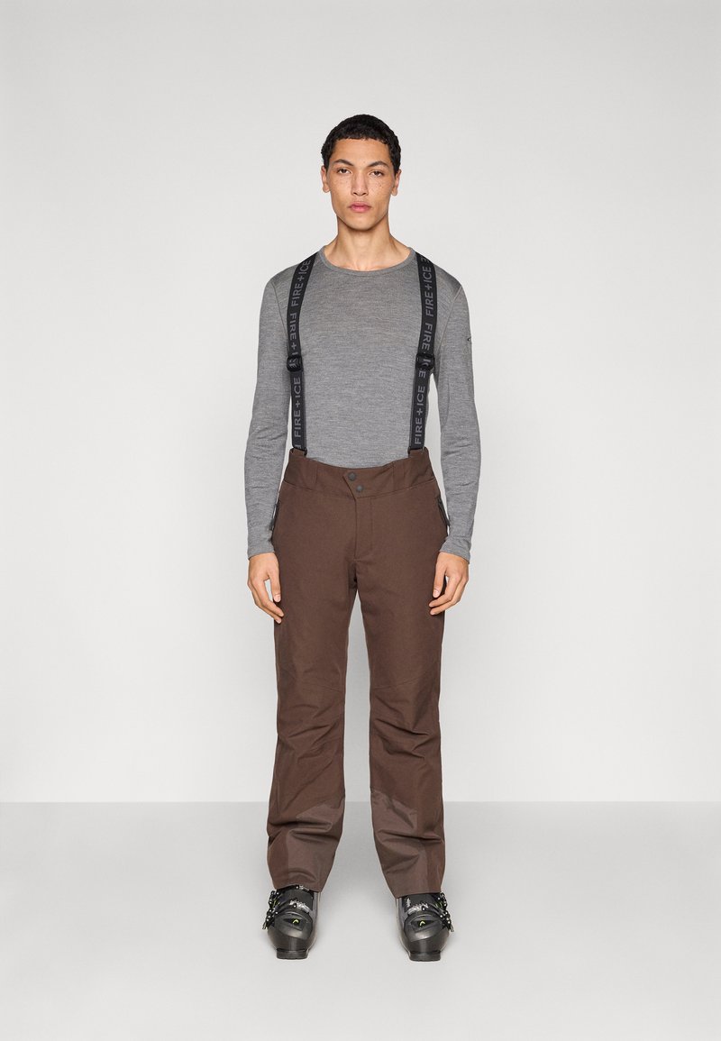 Καφέ παντελόνια σκι με ρυθμιζόμενες μαύρες τιράντες, που κλείνουν με κουμπί και έχουν φερμουάρ στα πλάγια, συνδυασμένα με μαύρα παπούτσια σκι και γκρι μπλούζα με μακριά μανίκια.