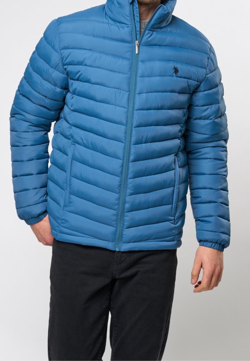 Homme portant une veste matelassée bleue zippée avec des poches latérales et un pantalon noir, debout devant un fond uni.