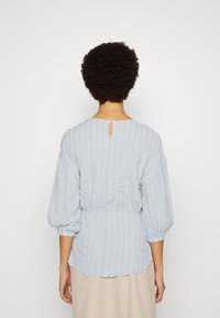 Blusa texturizada azul-claro com costas em forma de chave, mangas bufantes elásticas e cintura ajustada. Apresenta riscas verticais e bainha em forma de escalope.