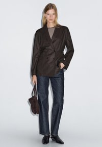 Brauner zweireihiger Blazer über einem gestreiften Oberteil, kombiniert mit dunklen geraden Jeans und schwarzen Loafers. Das Model hält eine braune Wildledertasche.