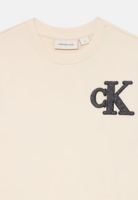 Krämfärgad sweatshirt med rundad halsringning, prydd med en svart glitter "ck"-logotyp på vänster bröst. Etikett visar storlek 8. Mjuk textur.
