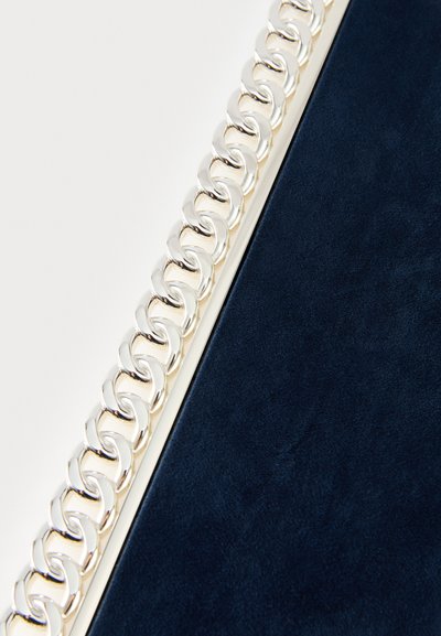 Sidabrinė grandinėlės detalė palei tamsiai mėlynos tekstūruotos zomšos paviršiaus kraštą. Grandinėlė sudaryta iš lygių, susikertančių blizgaus metalo jungčių.