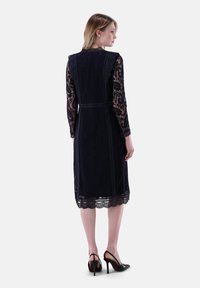 Navyblaues Spitzenkleid mit langen Ärmeln, gewellten Details und einem Taillengürtel. Verfügt über transparente Spitzelemente und einen knielangen Saum.