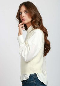 Maglione senza maniche color crema sopra una camicia bianca con colletto, con bordi a costine e un piccolo decoro sullo scollo.