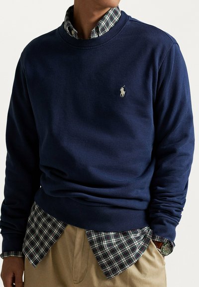 Felpa blu navy in misto cotone con collo a coste. Presenta un logo ricamato sul petto e una camicia a quadri sovrapposta sotto.