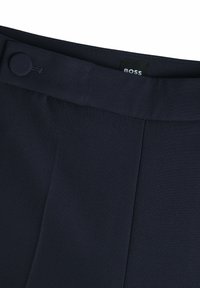 Marinblå byxor med rak front, prydliga sömmar, knappstängning och ett märke vid midjan. Tillverkade av strukturerad tyg.