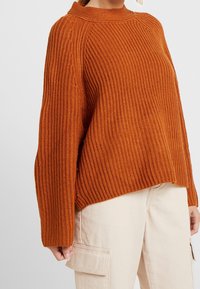 Kvinna klädd i en ribbstickad, bränd orange tröja med långa ärmar och beige cargobyxor med sido¬fickor, stående mot en enfärgad bakgrund.