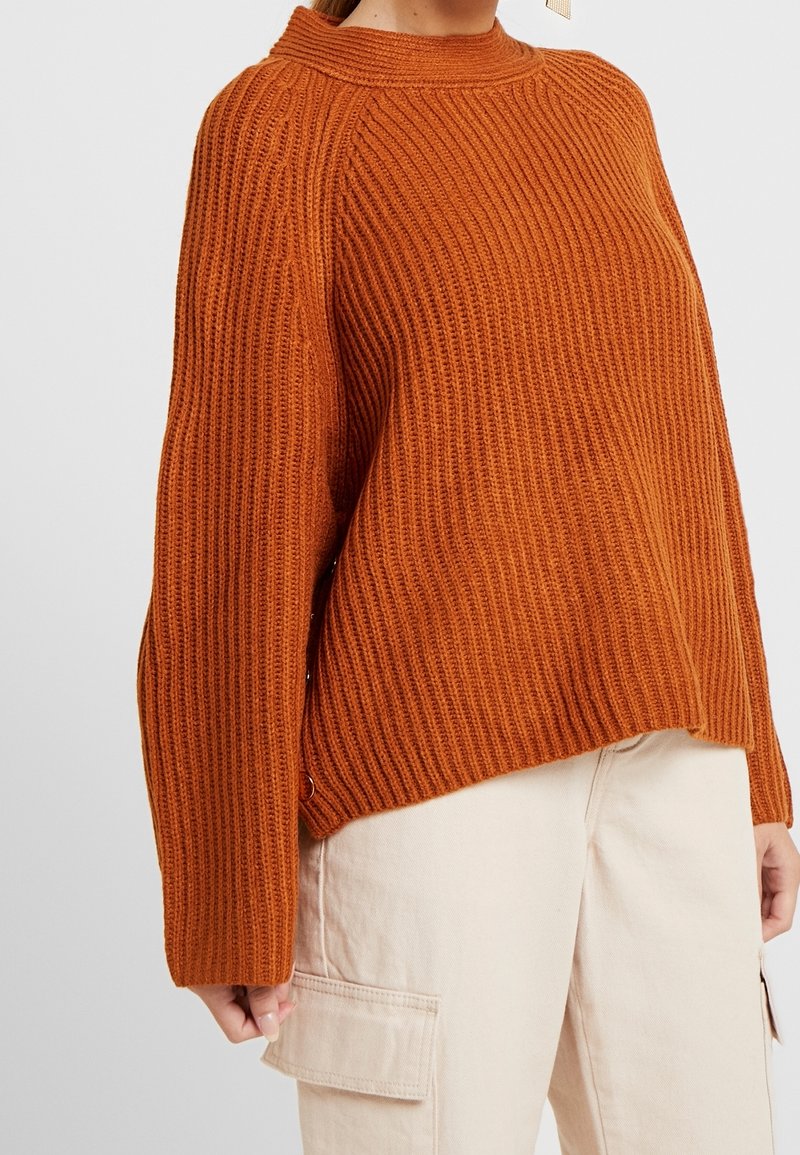 Nainen pukeutuneena poltettua oranssia ribbineulepaitaa pitkine hihoineen ja beigenvärisiin cargohousuihin sivutaskuilla, seisoo yksiväristä taustaa vasten.