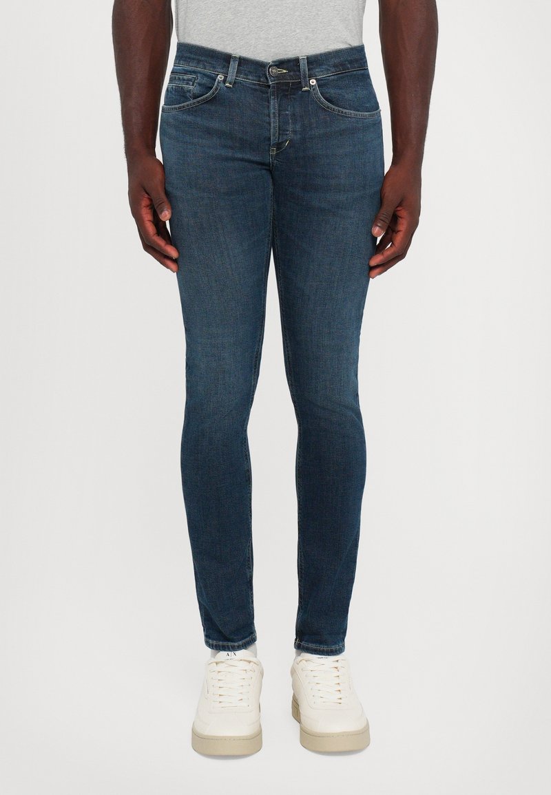 Homme portant un jean bleu foncé ajusté, des baskets blanches et une chemise gris clair, debout les mains détendues le long du corps.